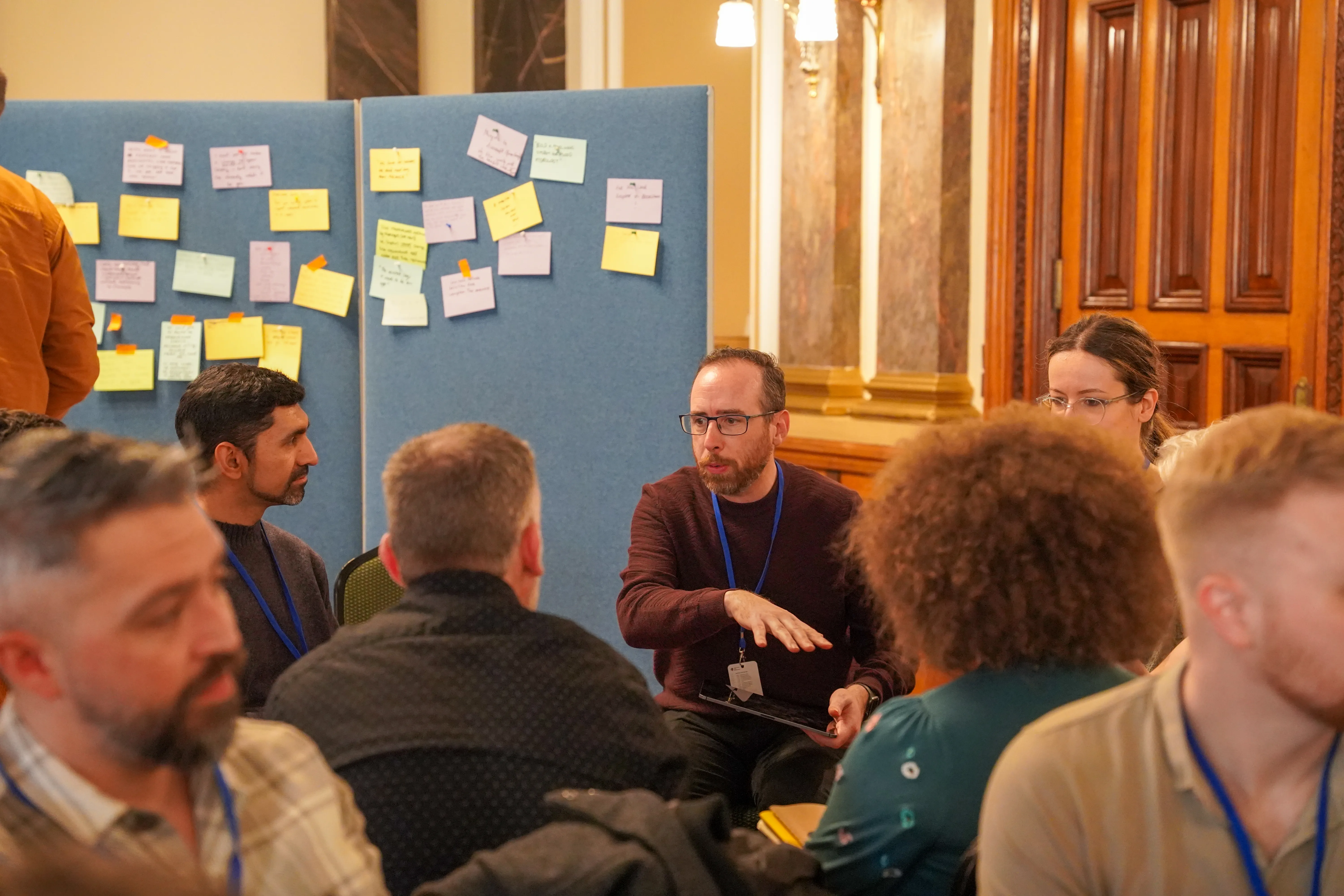 A large group of participants gathering in a spacious hall for a session. Behind them, a wall is covered in colorful sticky notes, highlighting a collaborative brainstorming process.